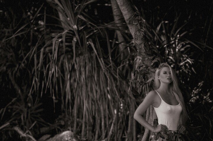 Blonde woman wearing a white top posing among dark tropical plants in a monochrome portrait