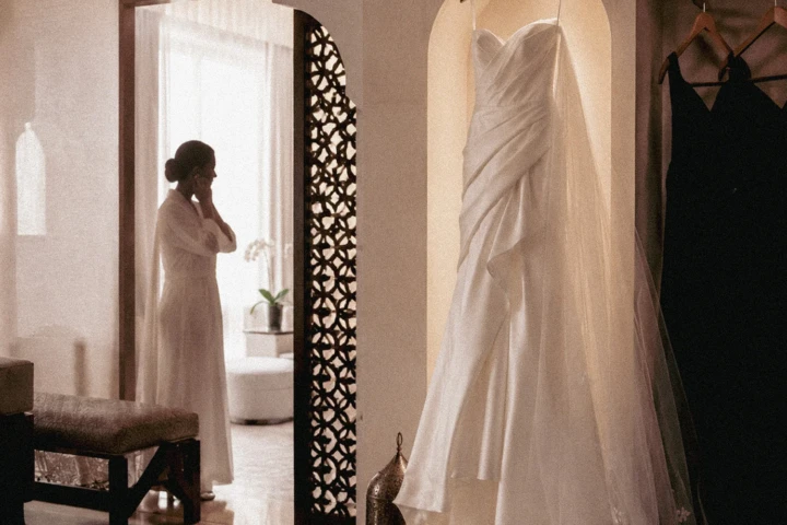 White wedding dress hanging in an elegant Abu Dhabi bridal suite while the bride prepares in the background.