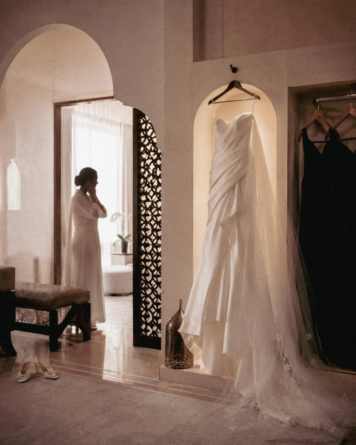 White wedding dress hanging in an elegant Abu Dhabi bridal suite while the bride prepares in the background.