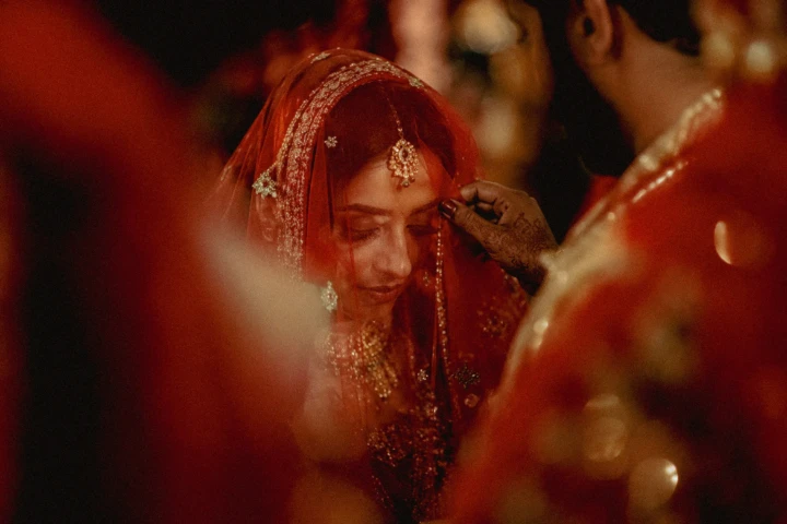 Afghan bride wearing a red embroidered veil while a family member adjusts her jewelry during a wedding ceremony in Dubai