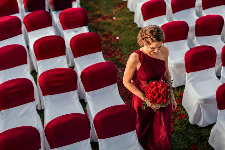 Bridesmaid carrying a red rose bouquet between burgundy and white chairs at an outdoor wedding ceremony in Dubai.