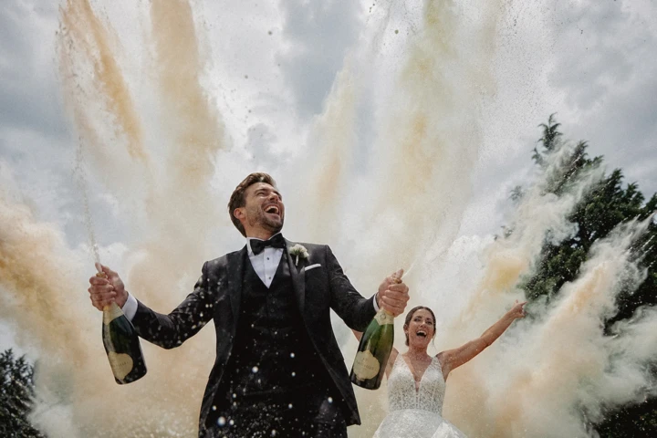 Laughing groom holding two champagne bottles with the bride celebrating behind him during an outdoor wedding.