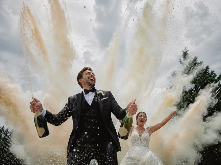 Laughing groom holding two champagne bottles with the bride celebrating behind him during an outdoor wedding.