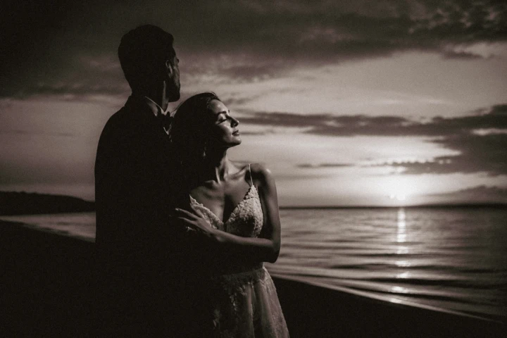 Bride and groom embracing beside the sea during a dramatic black-and-white sunset wedding portrait.