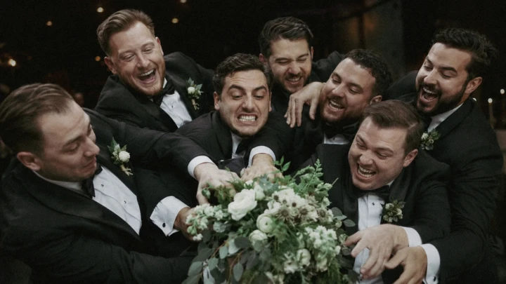 Groom and groomsmen in black tuxedos laughing and reaching for a white wedding bouquet in Istanbul.
