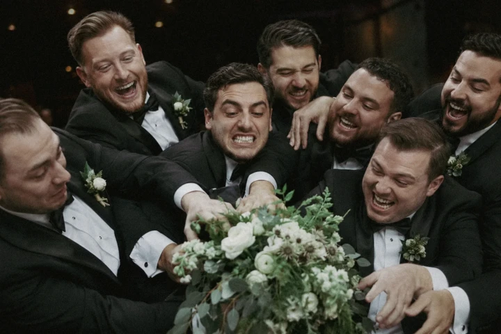 Groom and groomsmen in black tuxedos laughing and reaching for a white wedding bouquet in Istanbul.