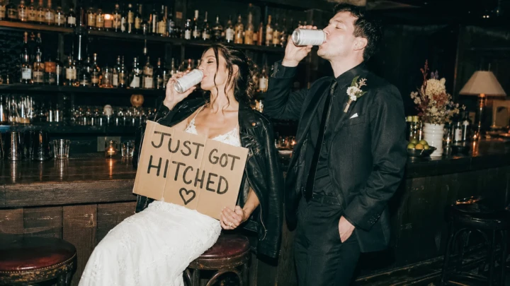 Bride and groom drinking together at a luxury hotel bar while holding a Just Got Hitched wedding sign.