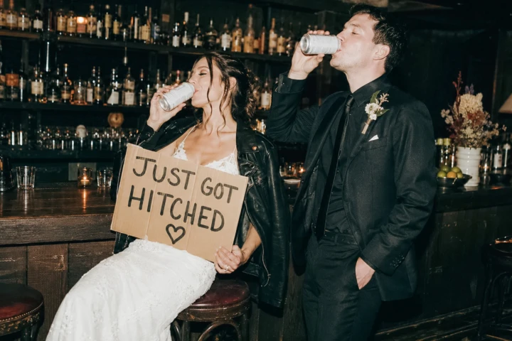 Bride and groom drinking together at a luxury hotel bar while holding a Just Got Hitched wedding sign.