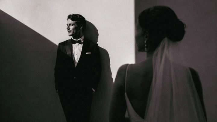 Groom standing in geometric sunlight while the bride appears softly out of focus in the foreground.