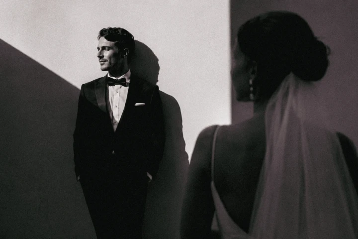 Groom standing in geometric sunlight while the bride appears softly out of focus in the foreground.