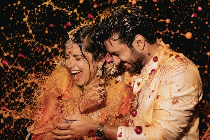 Indian bride and groom laughing as turmeric water and rose petals splash around them during a Haldi ceremony in Dubai.