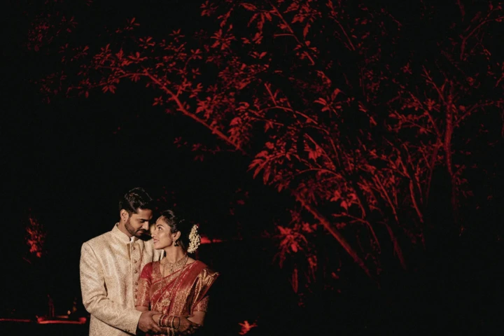 Indian bride and groom wearing traditional wedding attire beneath red illuminated trees at night in Dubai.