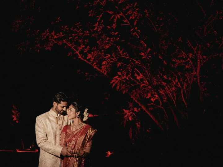 Indian bride and groom wearing traditional wedding attire beneath red illuminated trees at night in Dubai.