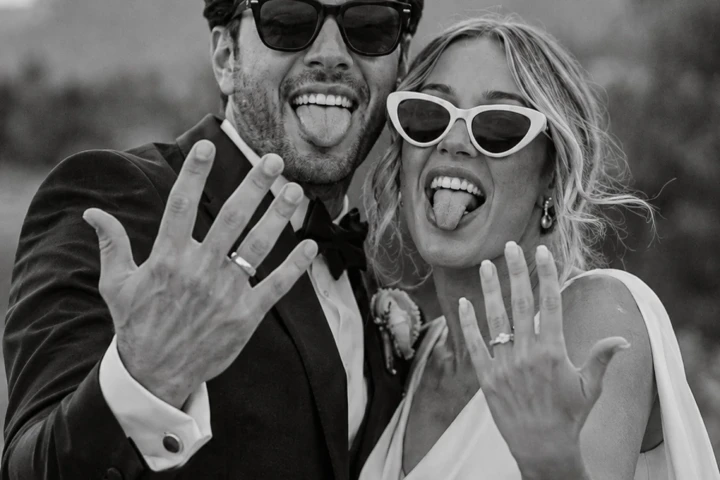 Bride and groom wearing sunglasses, showing their wedding rings and making playful expressions after the ceremony.