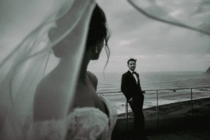 Bride in the foreground with her veil framing the groom beside the sea during a coastal wedding in Turkey.