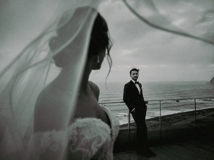 Bride in the foreground with her veil framing the groom beside the sea during a coastal wedding in Turkey.