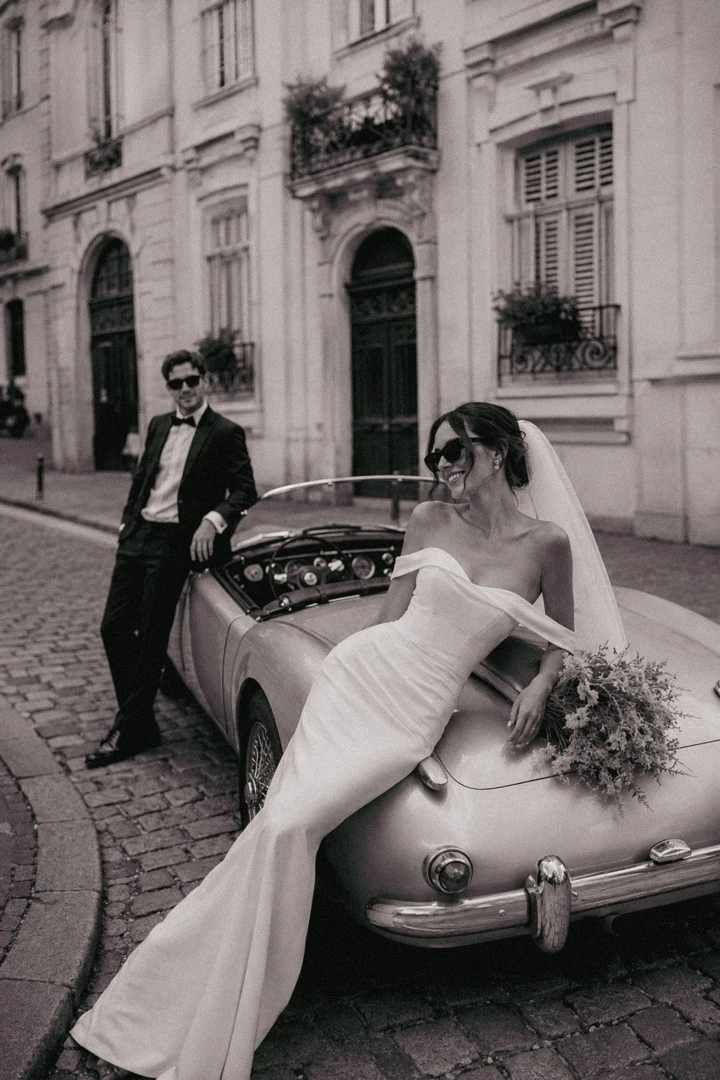 Bride sitting on a vintage convertible with the groom standing nearby on a historic cobblestone street in Turkey.