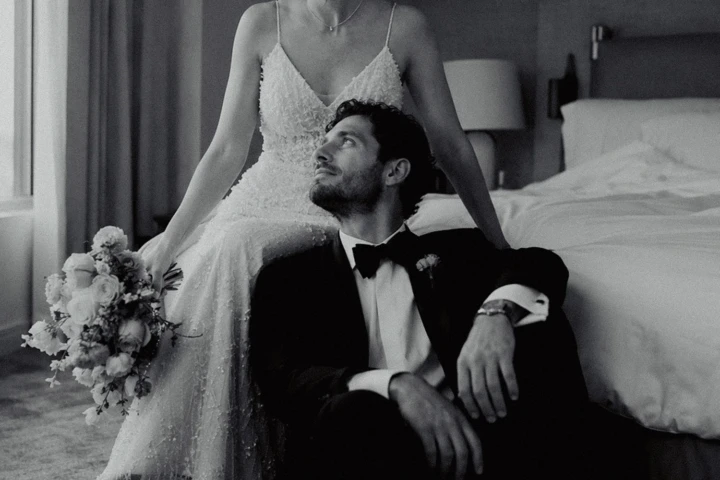 Bride seated beside a hotel window with her bouquet while the groom sits on the floor during a UAE wedding portrait.