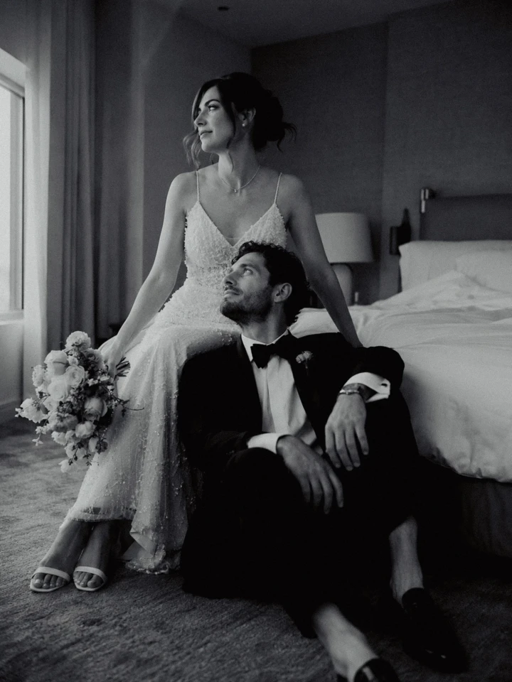 Bride seated beside a hotel window with her bouquet while the groom sits on the floor during a UAE wedding portrait.