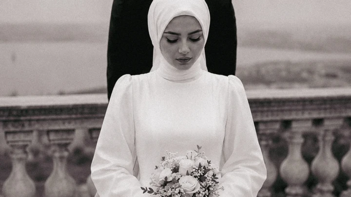 Muslim bride in a white hijab holding a floral bouquet with the groom standing behind her during a wedding portrait in the UAE.