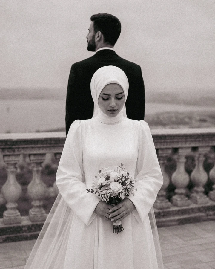 Muslim bride in a white hijab holding a floral bouquet with the groom standing behind her during a wedding portrait in the UAE.