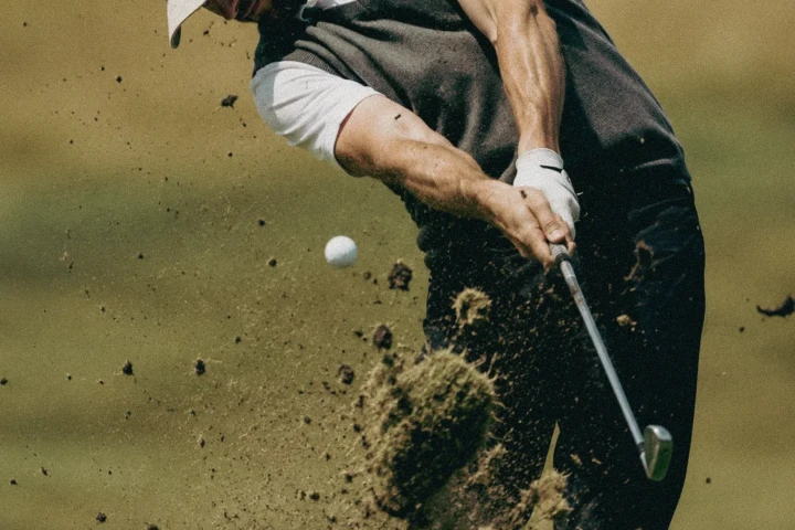 Golfer striking a ball with an iron as a large divot and soil fly through the air