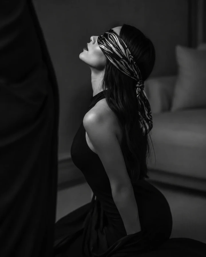 Black and white profile portrait of an adult woman wearing a patterned silk blindfold and dark dress in dramatic low-key light.
