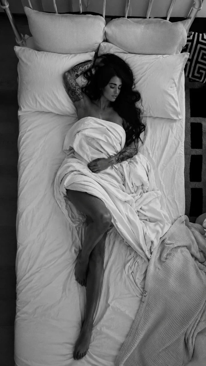 Overhead black and white portrait of an adult woman with tattoos lying in bed under white sheets in a relaxed bedroom setting.