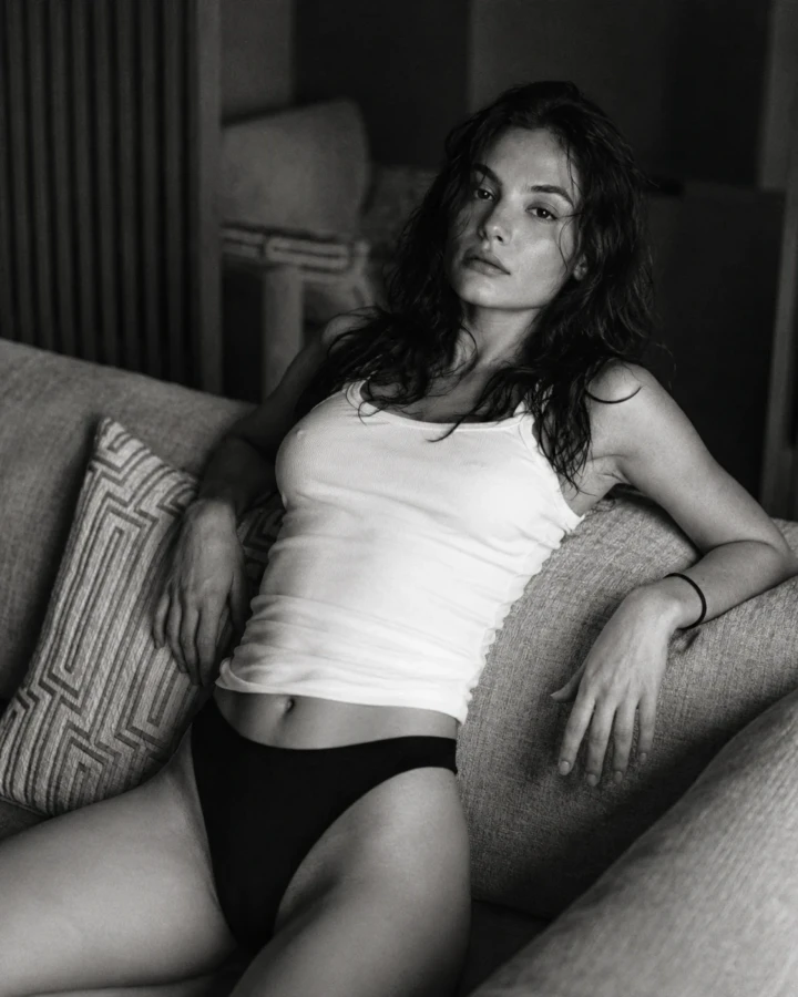 Black and white portrait of an adult woman in a tank top and briefs reclining casually on a sofa in a home interior.