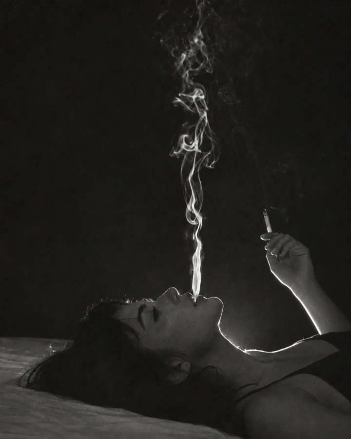 Black and white cinematic boudoir portrait of an adult woman reclining in dramatic low light with smoke rising above her face.