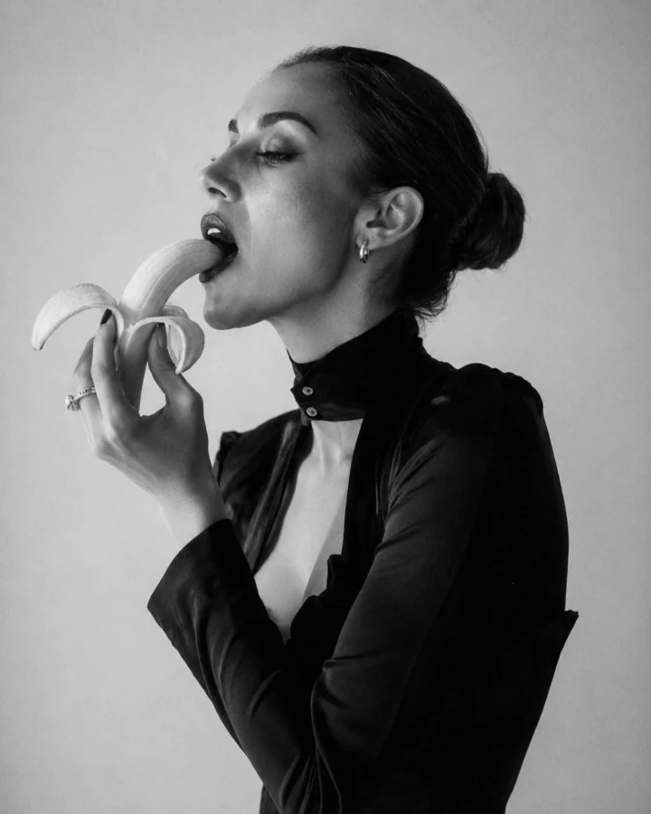 Black and white editorial profile of an adult woman in a dark fashion outfit holding a peeled banana against a minimal studio background.