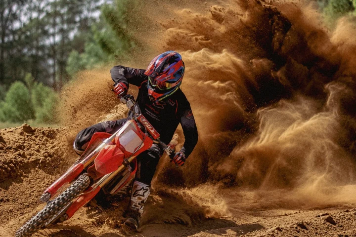 Motocross rider leaning a red dirt bike through sand with a large dust cloud behind