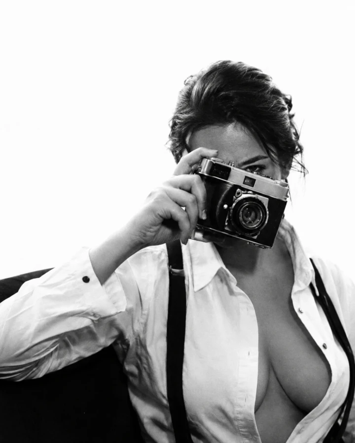 Black and white editorial portrait of an adult woman holding a vintage camera in front of her face while wearing a white shirt.