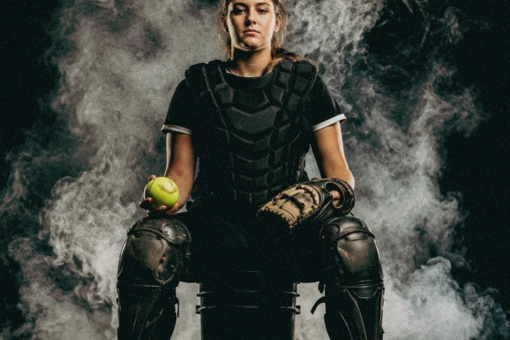 Female softball catcher wearing protective equipment and holding a ball in a smoky studio