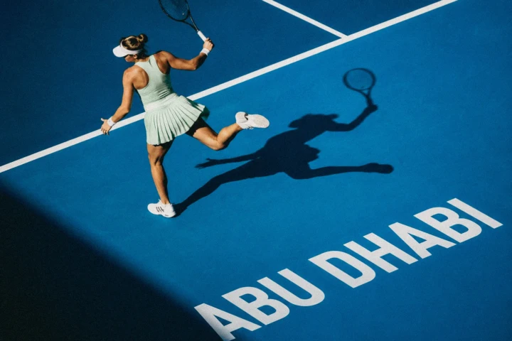 Female tennis player swinging a racket on a blue court marked Abu Dhabi with a long shadow