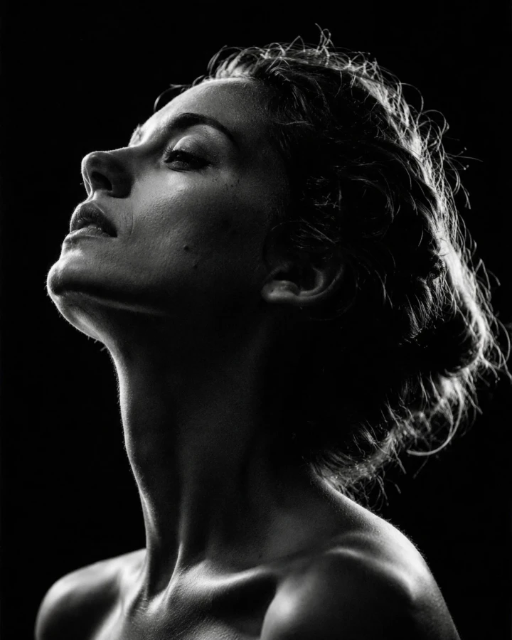 Dramatic black and white fine-art close-up portrait of an adult woman in profile with sculptural rim lighting against a dark background.