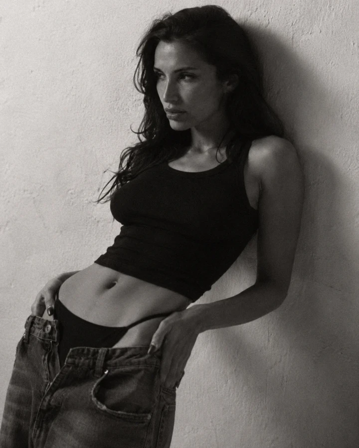 Black and white editorial portrait of an adult woman leaning against a textured wall in a fitted black top and relaxed denim jeans.