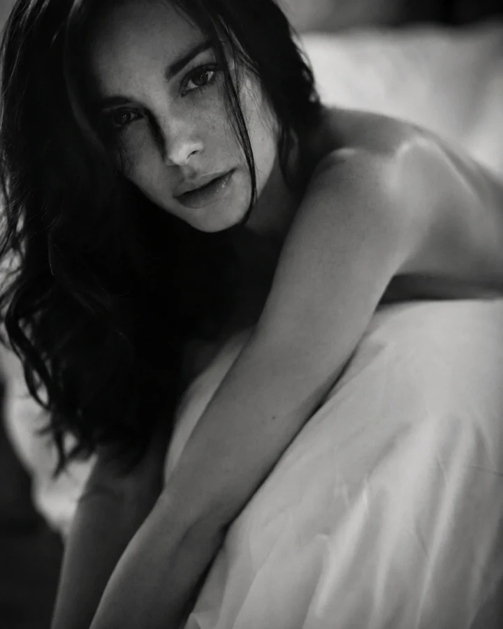 Close-up black and white portrait of an adult woman reclining on white bedding with natural skin texture and shallow depth of field.