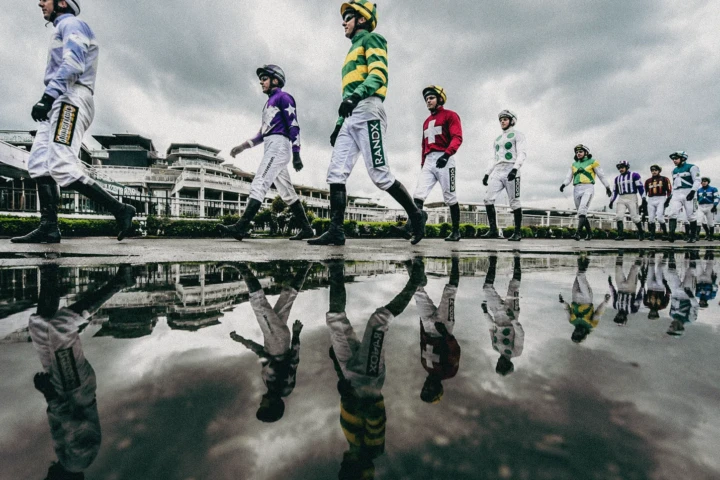 Group of jockeys walking beside a racecourse with their reflections visible in a puddle