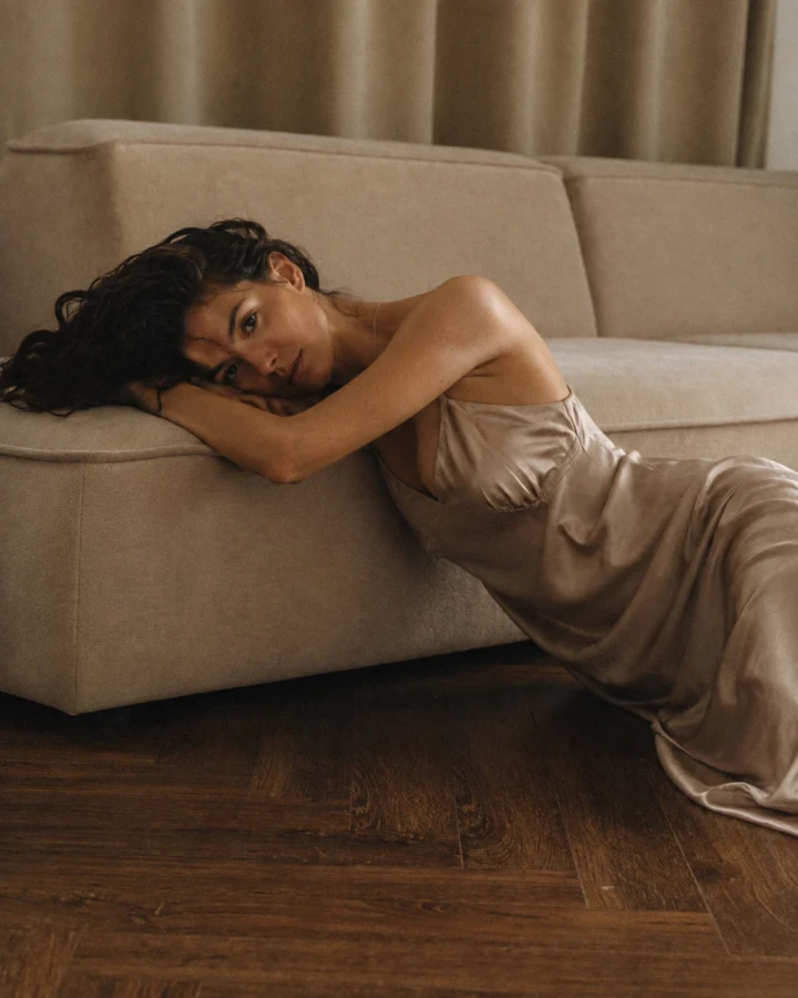 Elegant adult woman reclining beside a neutral sofa in a champagne satin dress during a warm natural-light editorial portrait.
