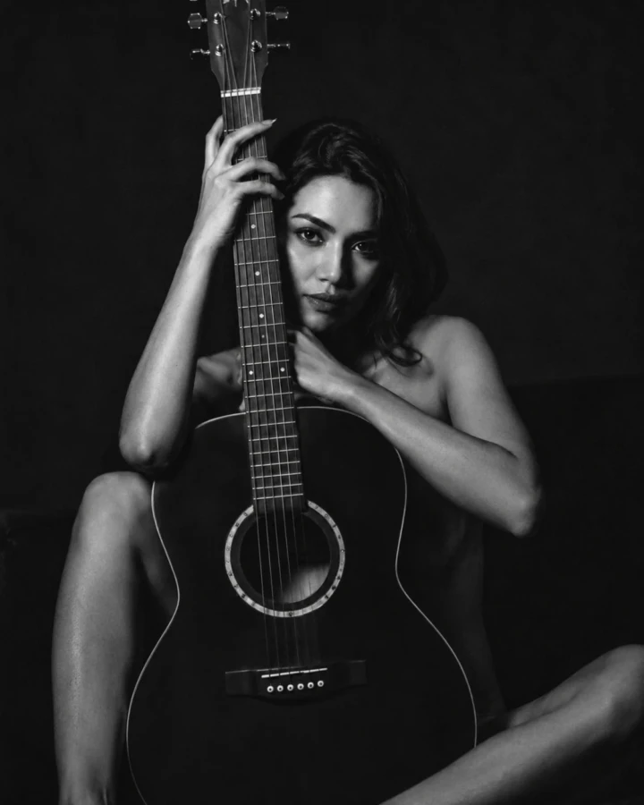 Black and white portrait of an adult woman posing behind an acoustic guitar in a dark intimate setting.
