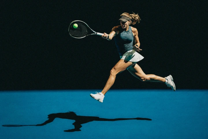 Female tennis player airborne while hitting a forehand on a blue court with a long shadow
