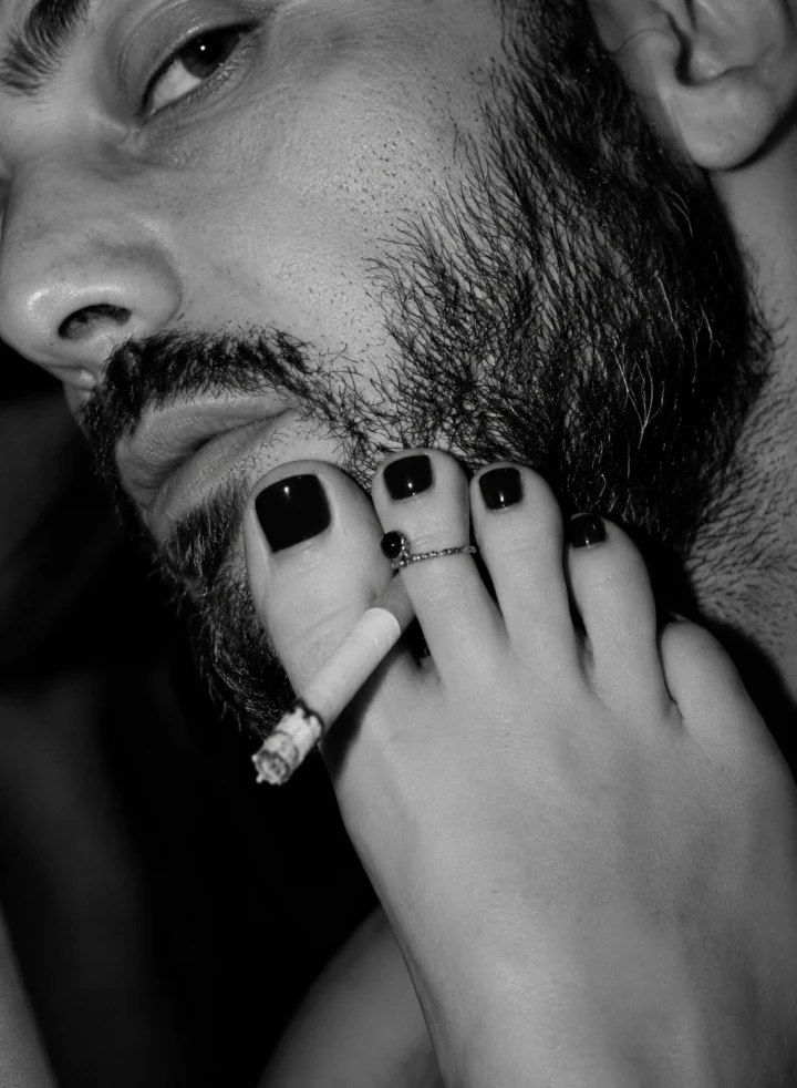 Close-up black and white editorial detail of a bearded man’s face with a woman’s foot, dark nail polish and a cigarette creating a provocative composition.