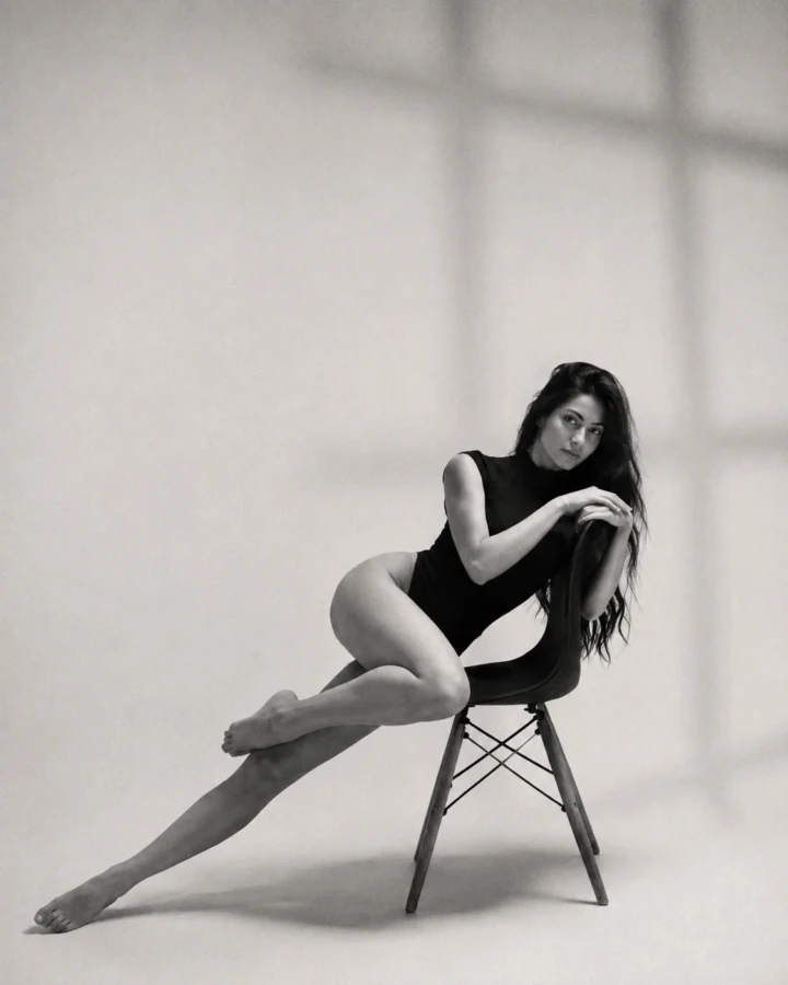 Minimal black and white studio portrait of an adult woman in a black bodysuit seated sideways on a chair against a plain background.