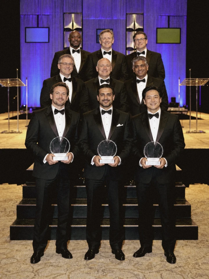 Formal group of corporate award recipients posing with trophies in front of a professionally lit event stage.