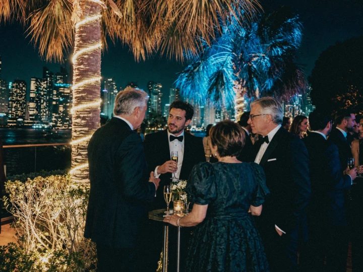 VIP guests networking at an elegant outdoor evening reception with a waterfront city skyline and illuminated palm trees.
